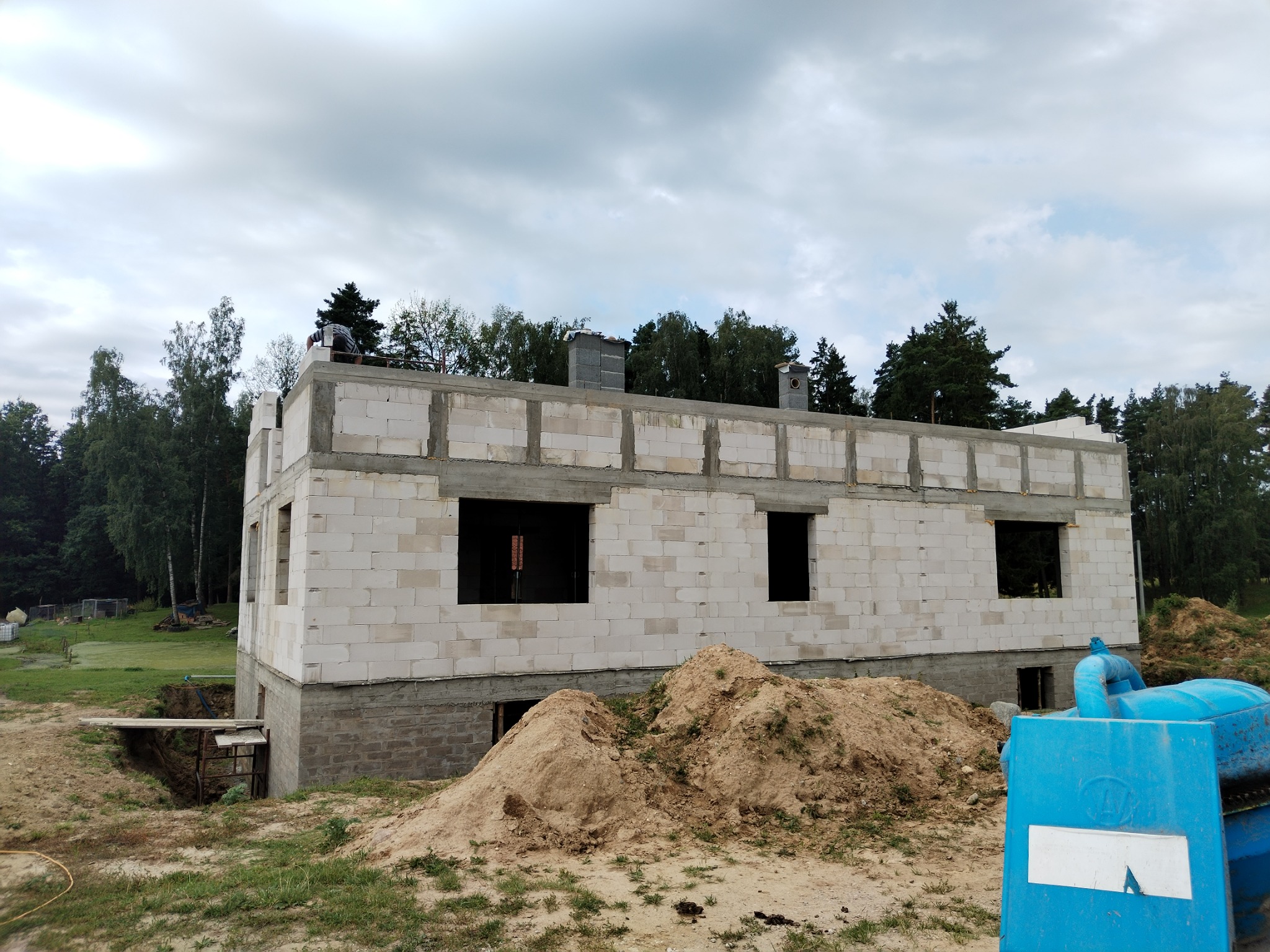 Budowany dom jednorodzinny z białych bloczków, z widocznymi fundamentami i otworami okiennymi, pracownik na stropie, kominy, hałdy piasku na pierwszym planie.