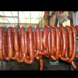 Wędzone kiełbasy zwisające z metalowego drążka w wędzarni, widoczny kubeł w tle.