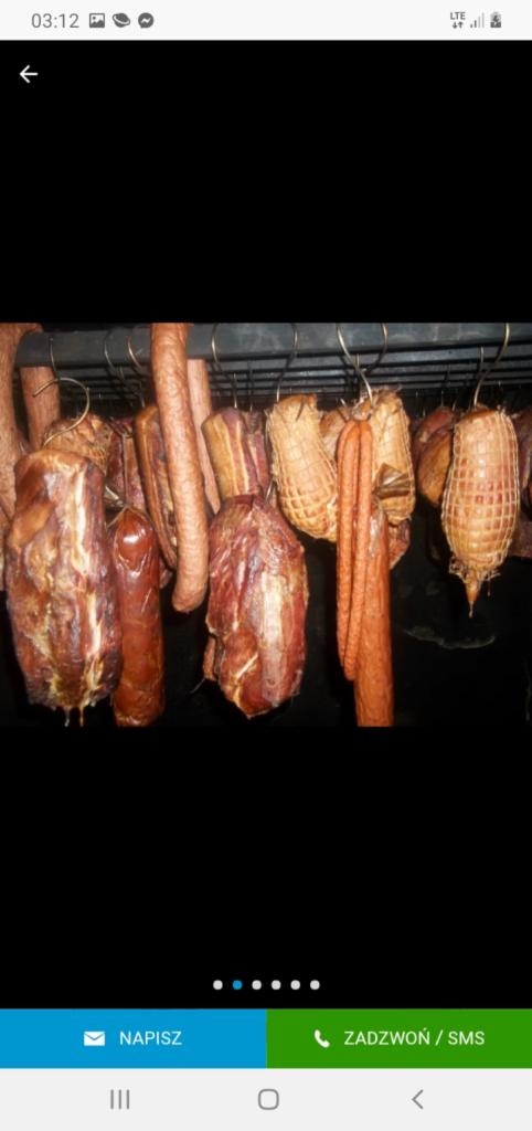 Wędzone wyroby wiszące na metalowych hakach w wędzarni, widoczne szynki, kiełbasy i balerony w siatkach wędliniarskich, oświetlenie punktowe.