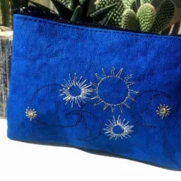 Niebieska kosmetyczka z motywem słońca i księżyca, wykonana haftem maszynowym w odcieniach srebra i złota, na tle kaktusa w drewnianej doniczce.