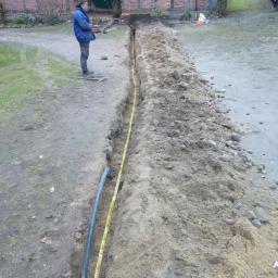 MM Malgorzata Skorska - Wąski wykop w ziemi z ułożonym kablem i taśmą ostrzegawczą, na tle starego budynku z cegły i pracownik nadzorujący.