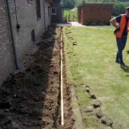 Wąski wykop wzdłuż ściany budynku z cegły, zabezpieczony żółtą taśmą ostrzegawczą, z fragmentami darni po jednej stronie i pracownikiem w pomarańczowej kamizelce w tle.