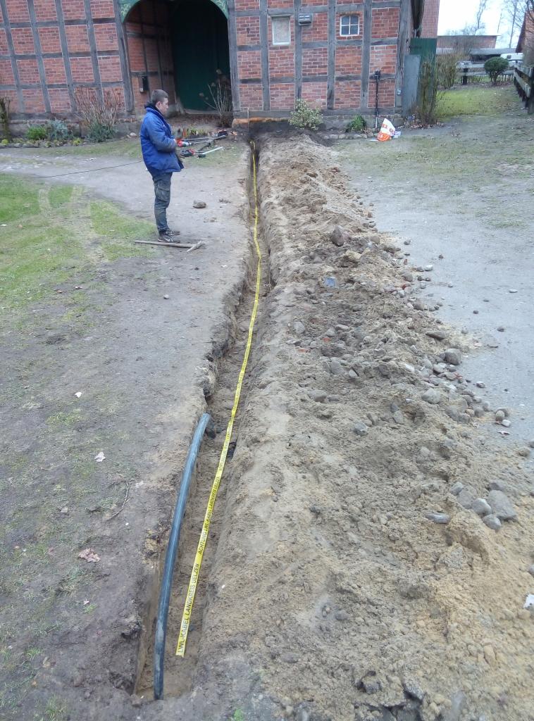 Wąski wykop w ziemi z ułożonym kablem i taśmą ostrzegawczą, na tle starego budynku z cegły i pracownik nadzorujący.