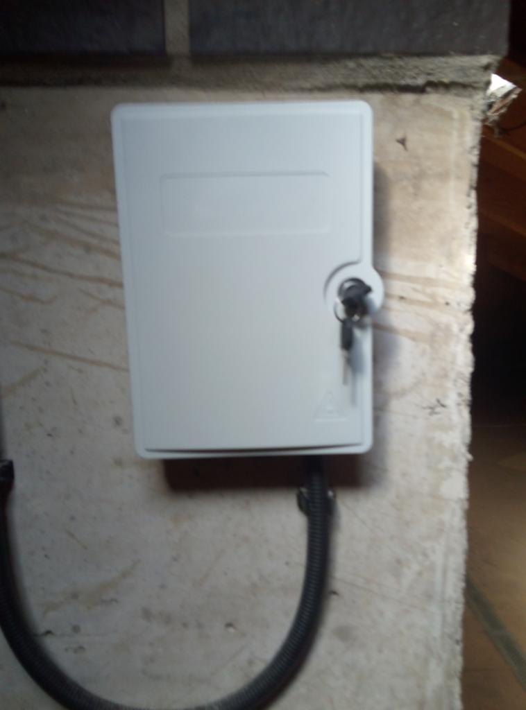 Zamknięta, biała skrzynka telekomunikacyjna z kluczykiem w zamku, zamontowana na surowej ścianie z widocznym przewodem ochronnym.