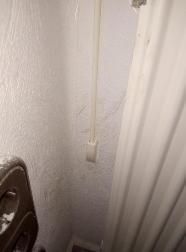 Biała rurka instalacji telekomunikacyjnej przymocowana do ściany z widoczną fakturą, obok grzejnik panelowy z widocznymi zabrudzeniami.