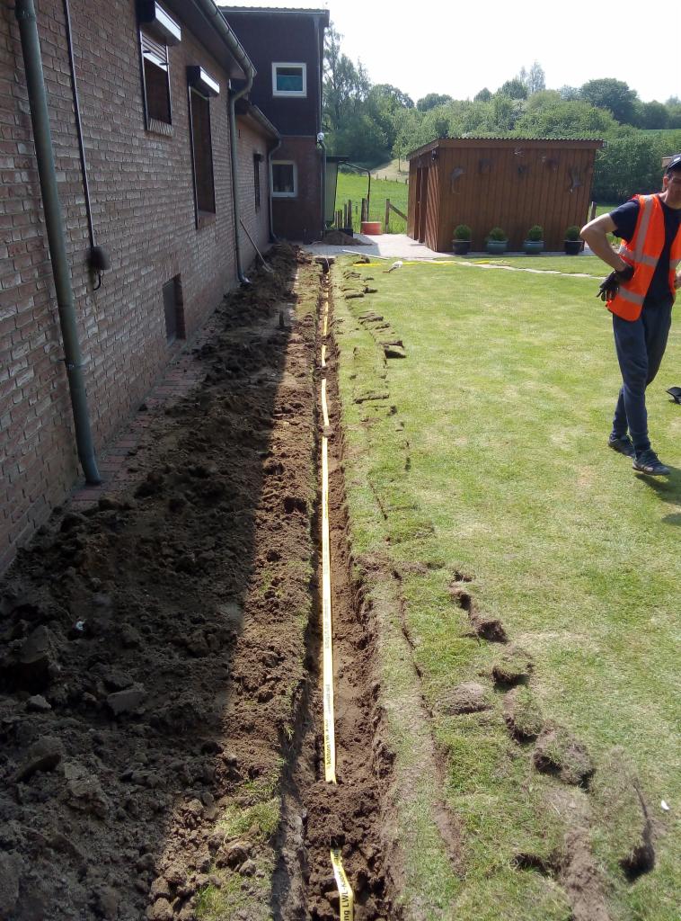 Wąski wykop wzdłuż ściany budynku z cegły, zabezpieczony żółtą taśmą ostrzegawczą, z fragmentami darni po jednej stronie i pracownikiem w pomarańczowej kamizelce w tle.
