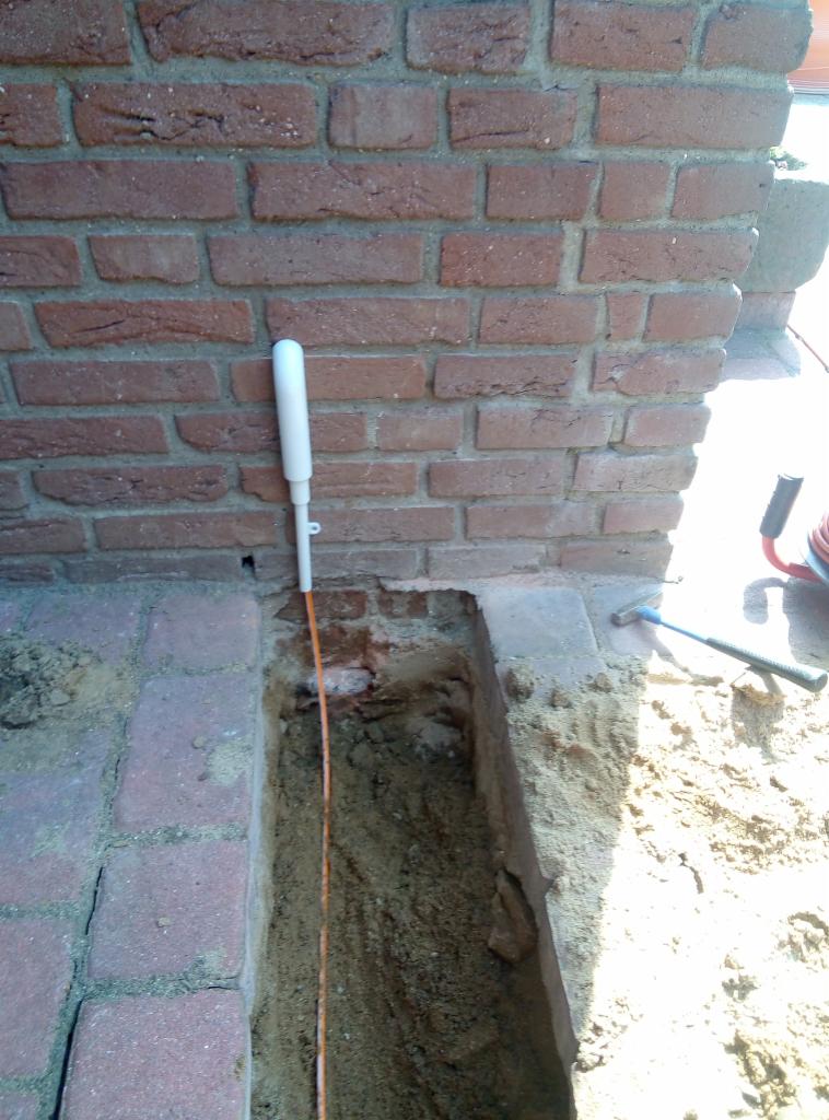 Wąski wykop wzdłuż ceglanej ściany budynku, z widocznym przewodem telekomunikacyjnym i narzędziem pomiarowym z białą obudową.