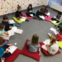Grupa dzieci siedzi na poduszkach w kręgu, trzymając w rękach kartki z obrazkami, na ścianie kolorowa tapeta w dłonie.