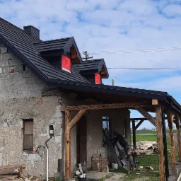 Dom w trakcie remontu z nowym, czarnym dachem z blachy na rąbek stojący, widoczne okna dachowe z folią ochronną i drewniana konstrukcja werandy z przodu budynku.