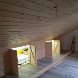 Adaptacja poddasza: drewniane zabudowy ze skrzydłowymi drzwiczkami w skosach, widoczna przestrzeń za drzwiczkami z żółtą ścianą i materacem, drewniana boazeria na ścianach i suficie, panele...