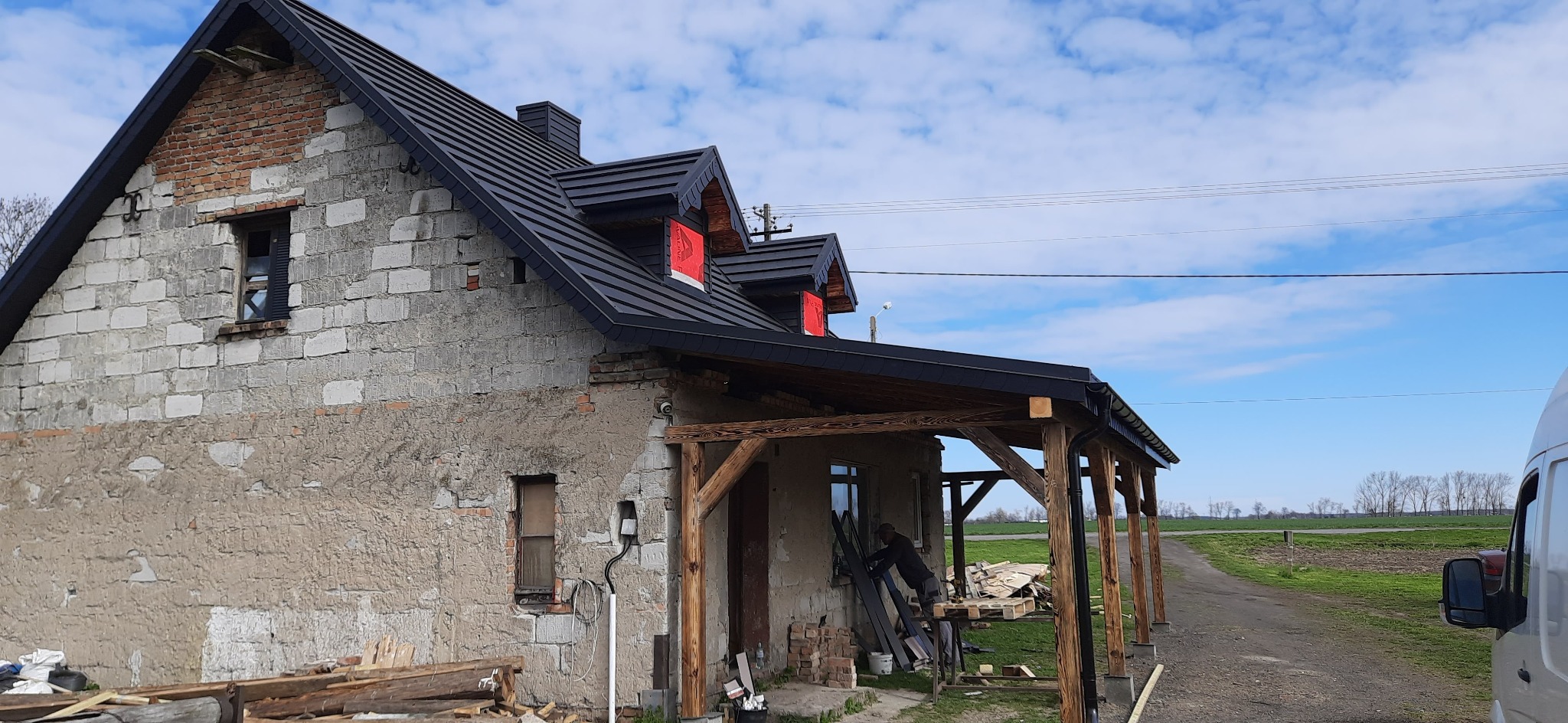 Dom w trakcie remontu z nowym, czarnym dachem z blachy na rąbek stojący, widoczne okna dachowe z folią ochronną i drewniana konstrukcja werandy z przodu budynku.