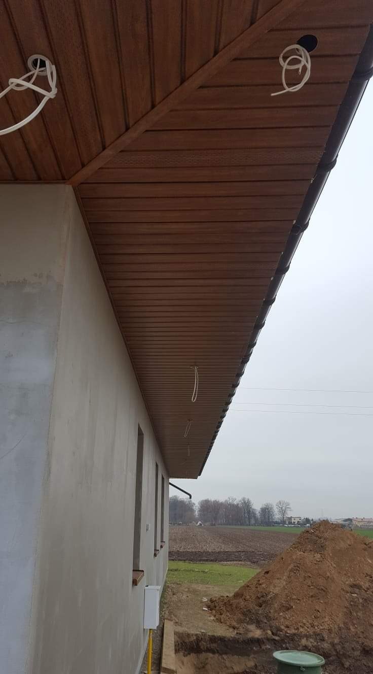 Drewniana podbitka dachowa w kolorze brązowym, widoczna z zewnątrz budynku, z wystającymi przewodami elektrycznymi, zamontowana przy ścianie budynku w trakcie prac wykończeniowych.