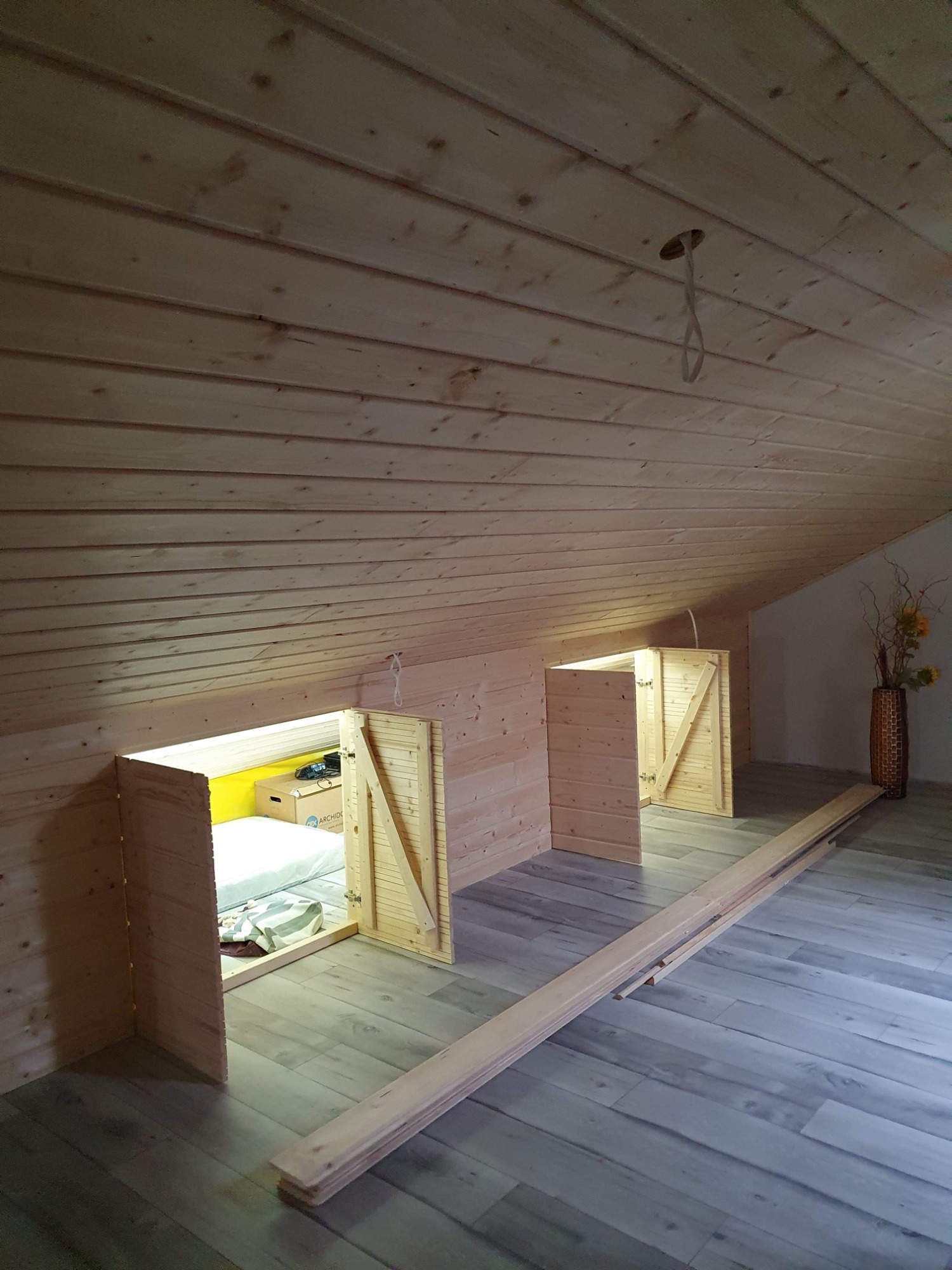 Adaptacja poddasza: drewniane zabudowy ze skrzydłowymi drzwiczkami w skosach, widoczna przestrzeń za drzwiczkami z żółtą ścianą i materacem, drewniana boazeria na ścianach i suficie, panele...