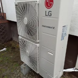 Zewnętrzna jednostka pompy ciepła LG Therma V, umieszczona na betonowych bloczkach, z widocznymi dwoma wentylatorami i kroplami deszczu na obudowie.