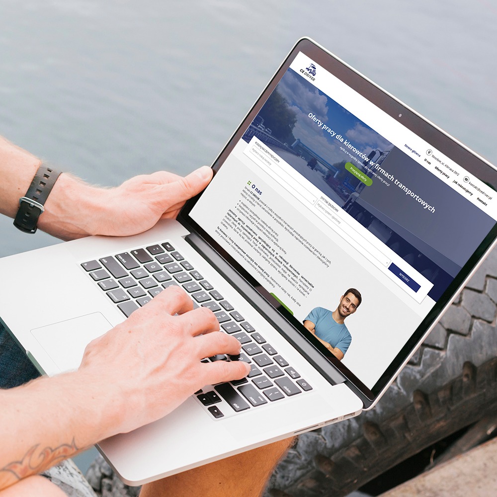Mężczyzna używa laptopa prezentującego stronę internetową firmy rekrutacyjnej dla kierowców, z widocznym logo CE DRIVER i ofertami pracy, na tle błękitnej wody i opony.