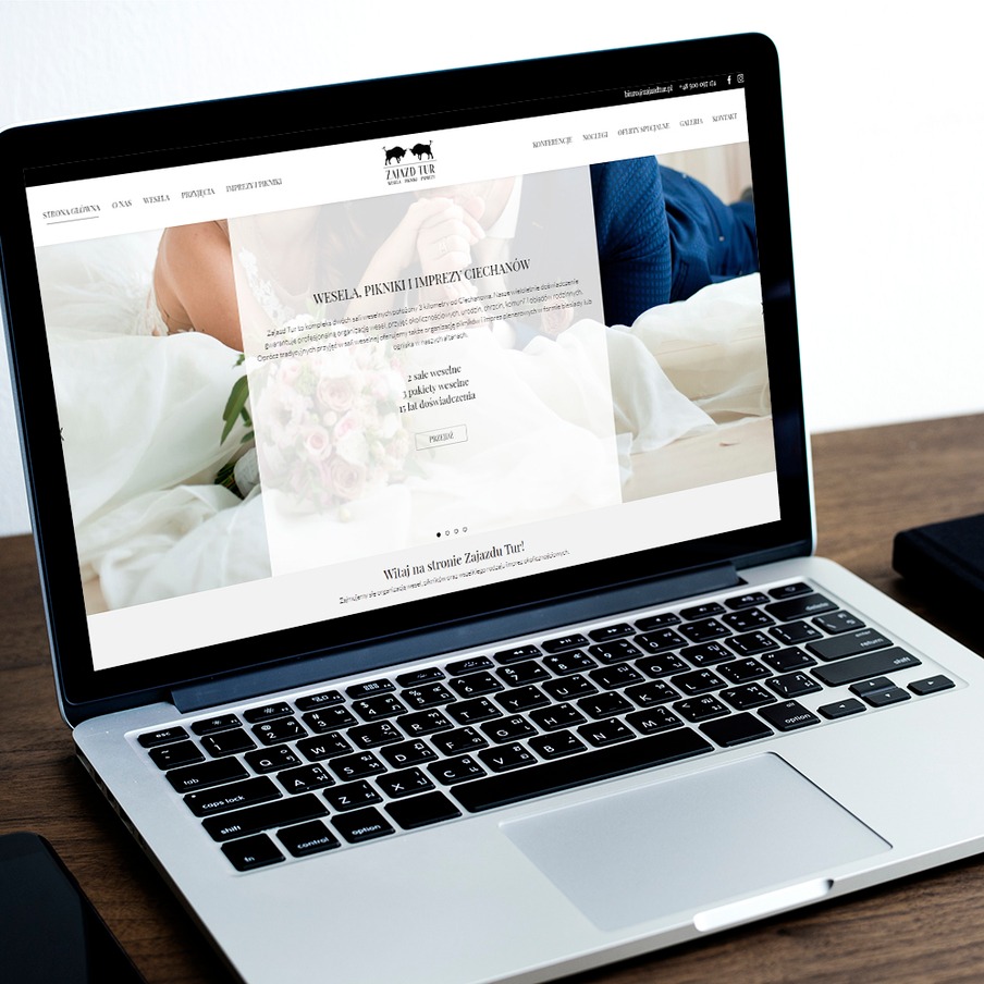 Ekran laptopa wyświetlający stronę internetową Zajazdu Tur z ofertą wesel, pikników i imprez w Ciechanowie, na drewnianym biurku obok leży smartfon i czarny notatnik.