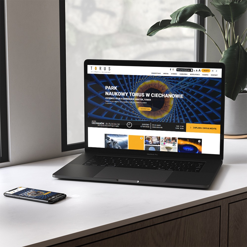 Laptop MacBook Pro prezentuje stronę internetową Parku Nauki Torus w Ciechanowie; identyczna strona widoczna na smartfonie obok, roślina doniczkowa w tle.