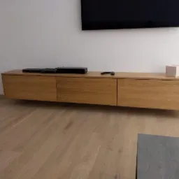 Drewniana szafka RTV wisząca na ścianie w minimalistycznym salonie, z widocznymi urządzeniami elektronicznymi i pilotem na blacie, na tle jasnej ściany i drewnianej podłogi.