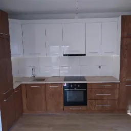 Nowoczesna kuchnia z białymi szafkami górnymi i drewnianymi szafkami dolnymi, z widocznym piekarnikiem i płytą grzewczą, oświetlona listwą LED pod szafkami wiszącymi.