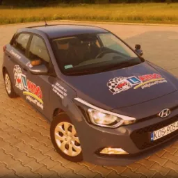 Szary samochód Hyundai z naklejkami firmy na parkingu z kostki brukowej, widziany z góry pod kątem w ciepłym świetle słonecznym.