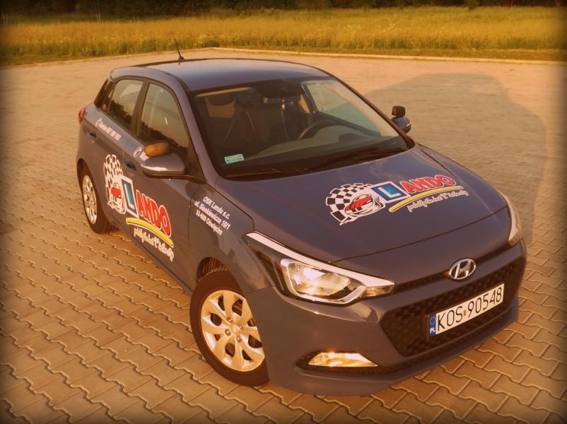 Szary samochód Hyundai z naklejkami firmy na parkingu z kostki brukowej, widziany z góry pod kątem w ciepłym świetle słonecznym.