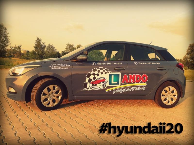 Szary Hyundai i20 z naklejkami z logo i numerami telefonów na boku, zaparkowany na chodniku z kostki brukowej, w tle zieleń.