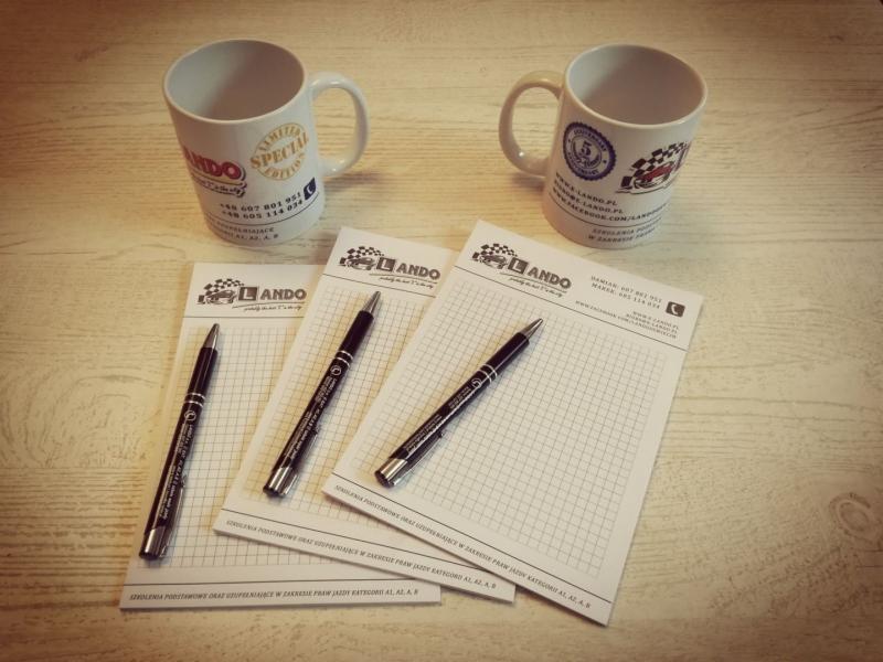 Dwa kubki z logo i danymi kontaktowymi, notatniki z kratką i długopisy na jasnym drewnianym stole, materiały promocyjne firmy.