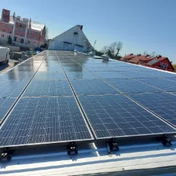 Rząd paneli słonecznych na metalowym dachu z widocznymi mocowaniami, w tle budynki mieszkalne i błękitne niebo.