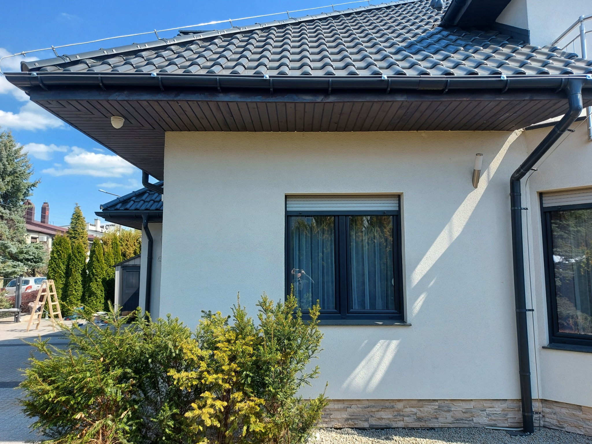 Zewnętrzny widok domu z zainstalowanym systemem odgromowym, widoczne elementy zabezpieczające na dachu i przewodzie odprowadzającym wzdłuż ściany, na tle błękitnego nieba.