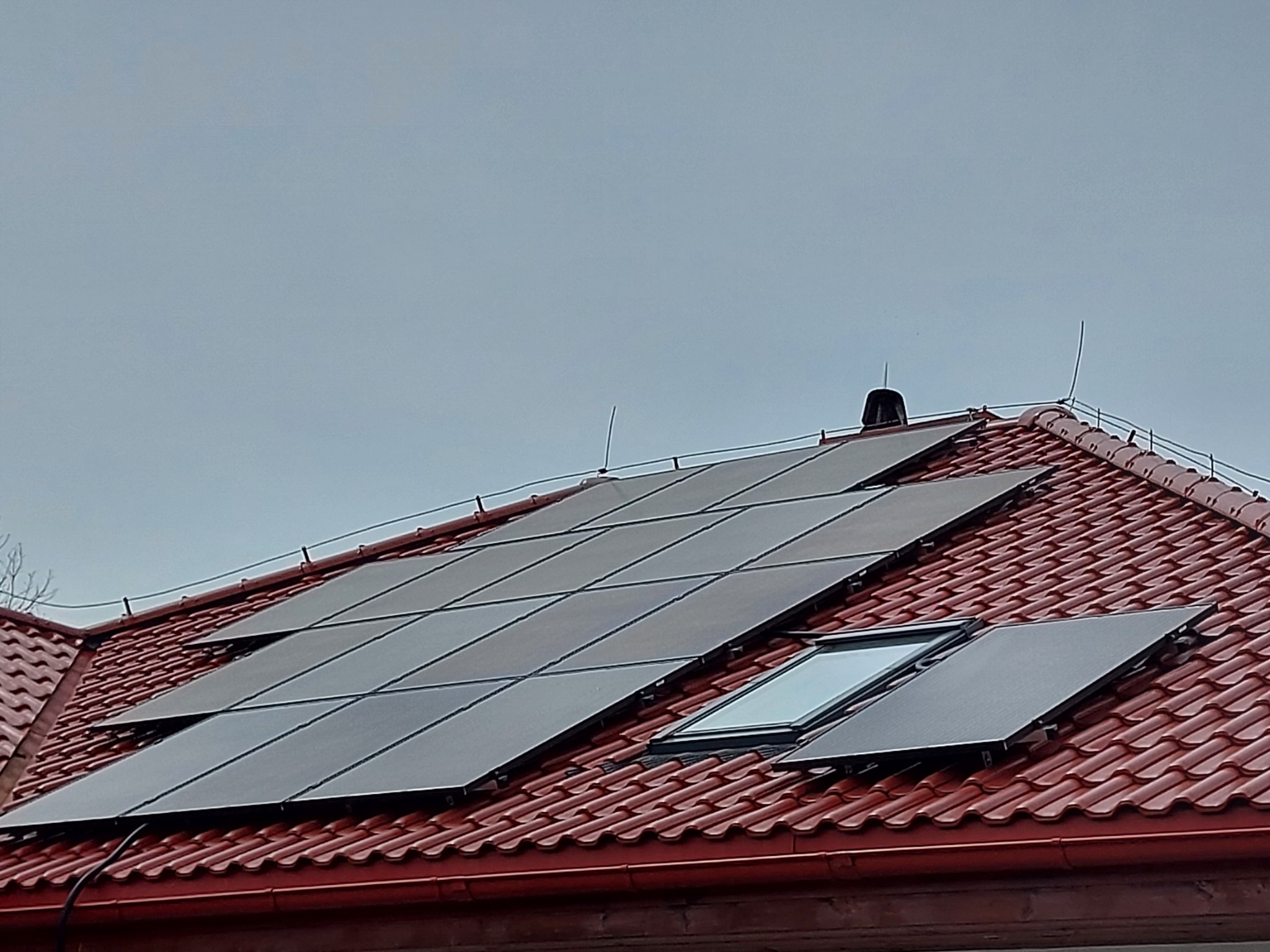 Czerwony dach pokryty panelami słonecznymi, widoczne okno dachowe i zabezpieczenie odgromowe na krawędzi dachu, pochmurne niebo w tle.