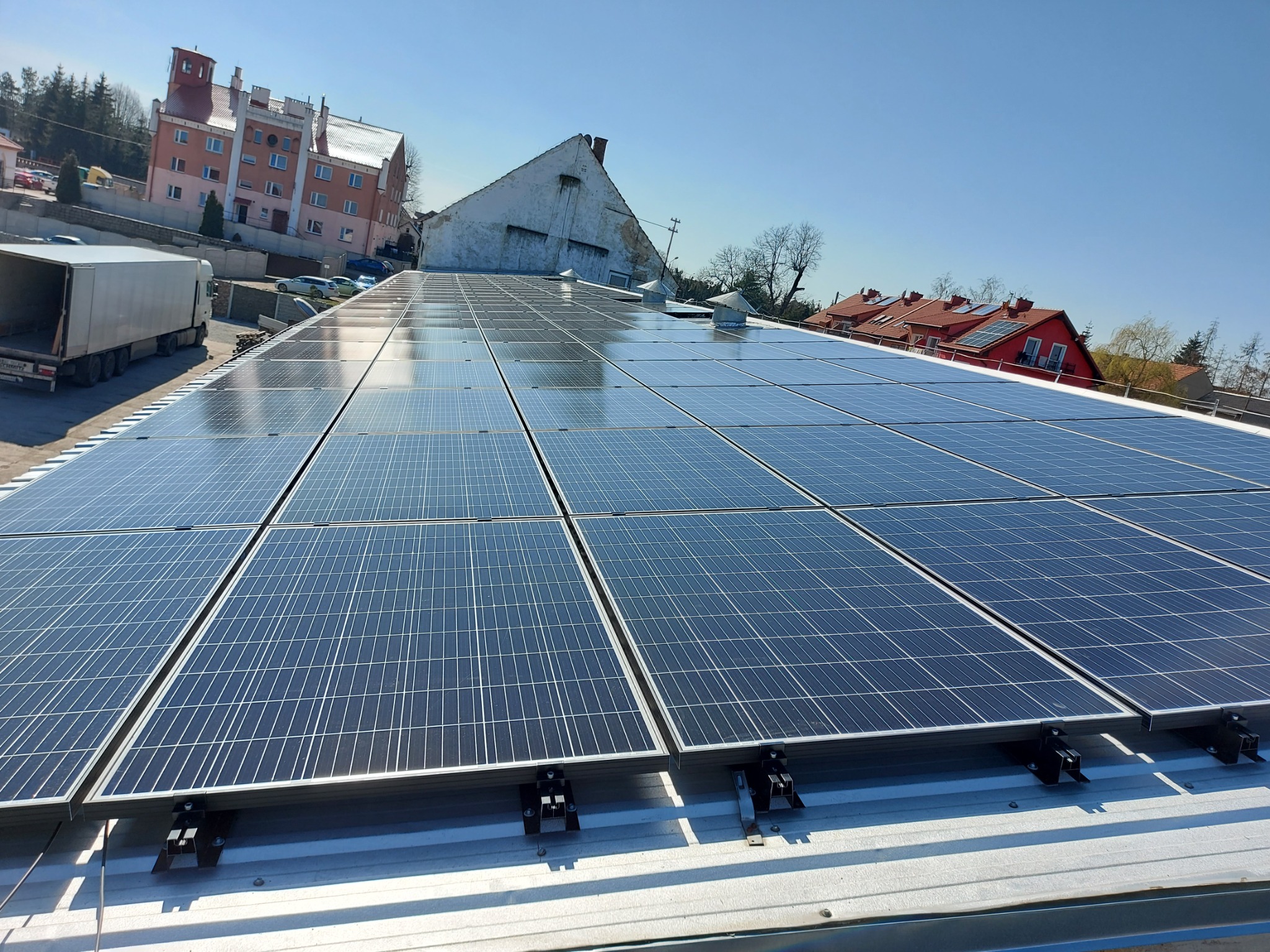 Rząd paneli słonecznych na metalowym dachu z widocznymi mocowaniami, w tle budynki mieszkalne i błękitne niebo.