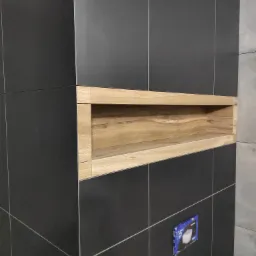 Czarne płytki ceramiczne z drewnianą wnęką w łazience, widoczna instalacja podtynkowa WC.