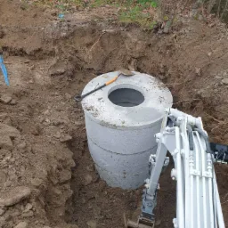 Montaż betonowego zbiornika w wykopie ziemnym przy użyciu minikoparki. Na górze leży łopata. Widok z góry na dół, oświetlenie dzienne.