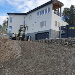 Nierówny teren przed nowoczesnym domem z szarą elewacją i garażem. Widoczny sprzęt budowlany i kontener. Częściowo ukończona elewacja. Błękitne niebo.