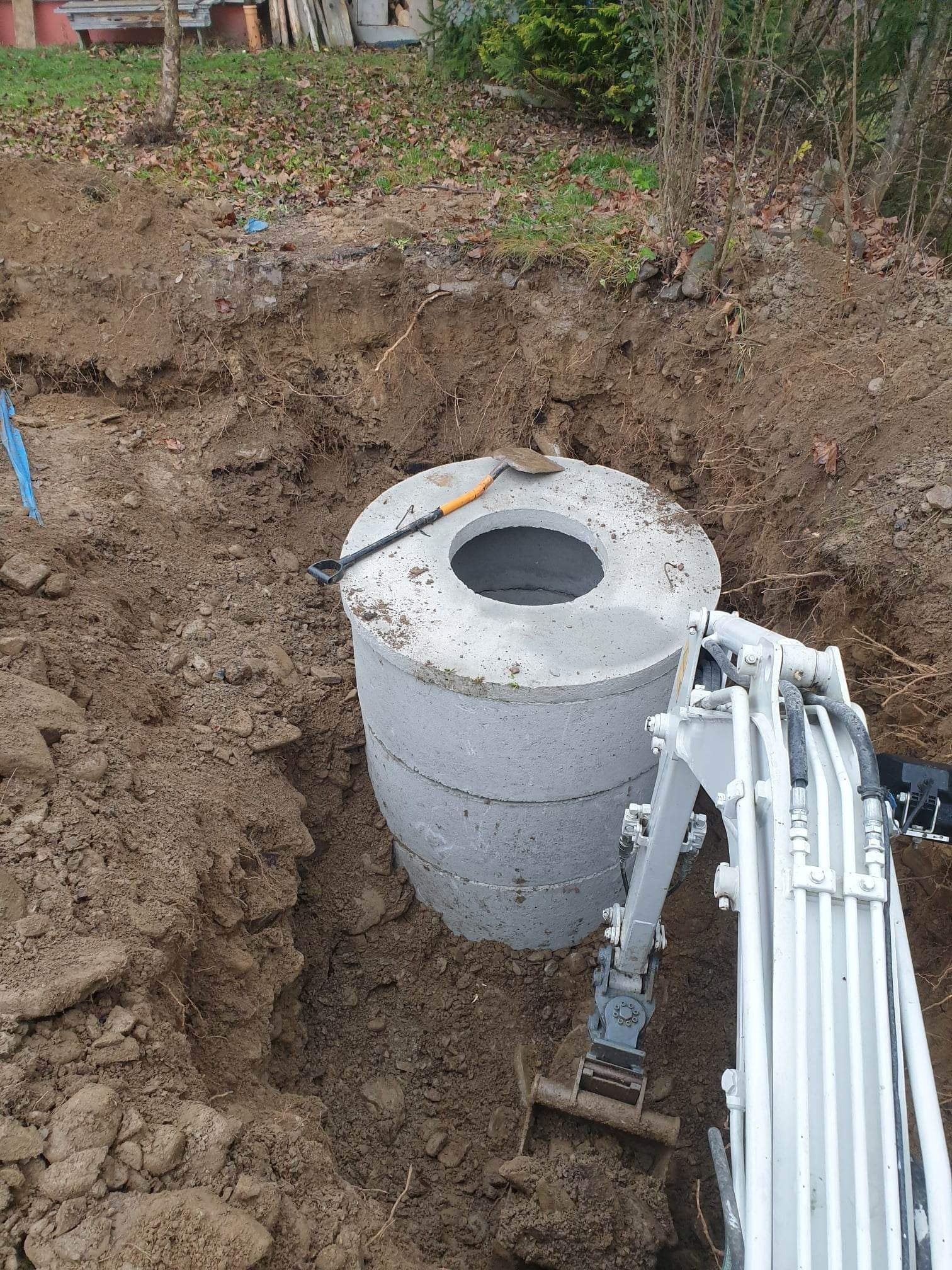 Montaż betonowego zbiornika w wykopie ziemnym przy użyciu minikoparki. Na górze leży łopata. Widok z góry na dół, oświetlenie dzienne.