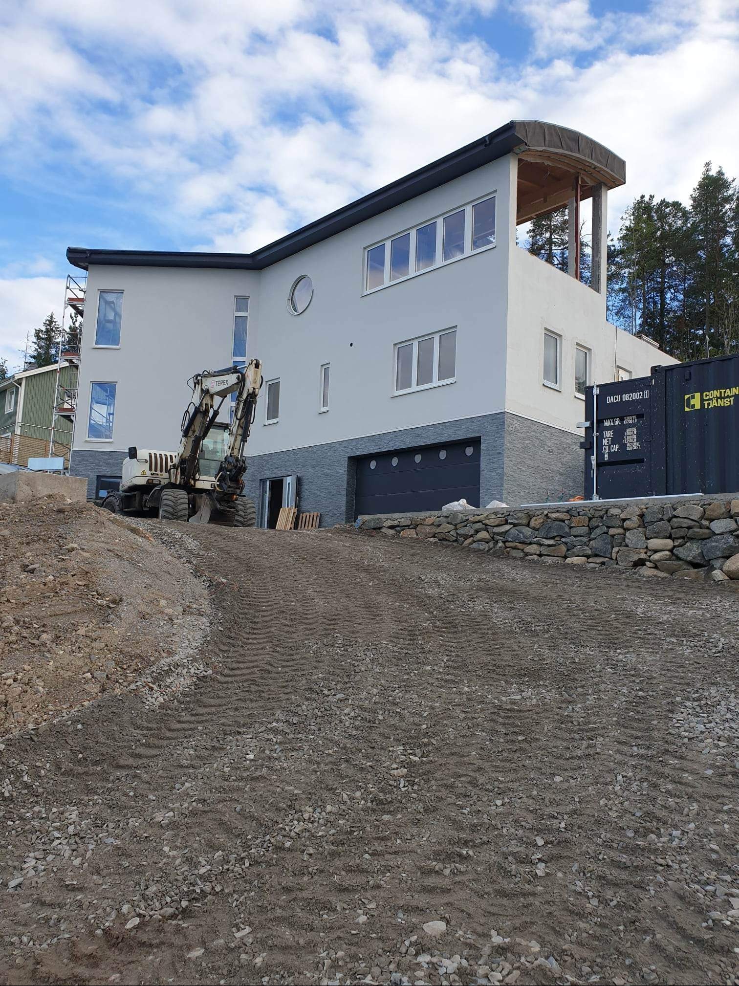 Nierówny teren przed nowoczesnym domem z szarą elewacją i garażem. Widoczny sprzęt budowlany i kontener. Częściowo ukończona elewacja. Błękitne niebo.