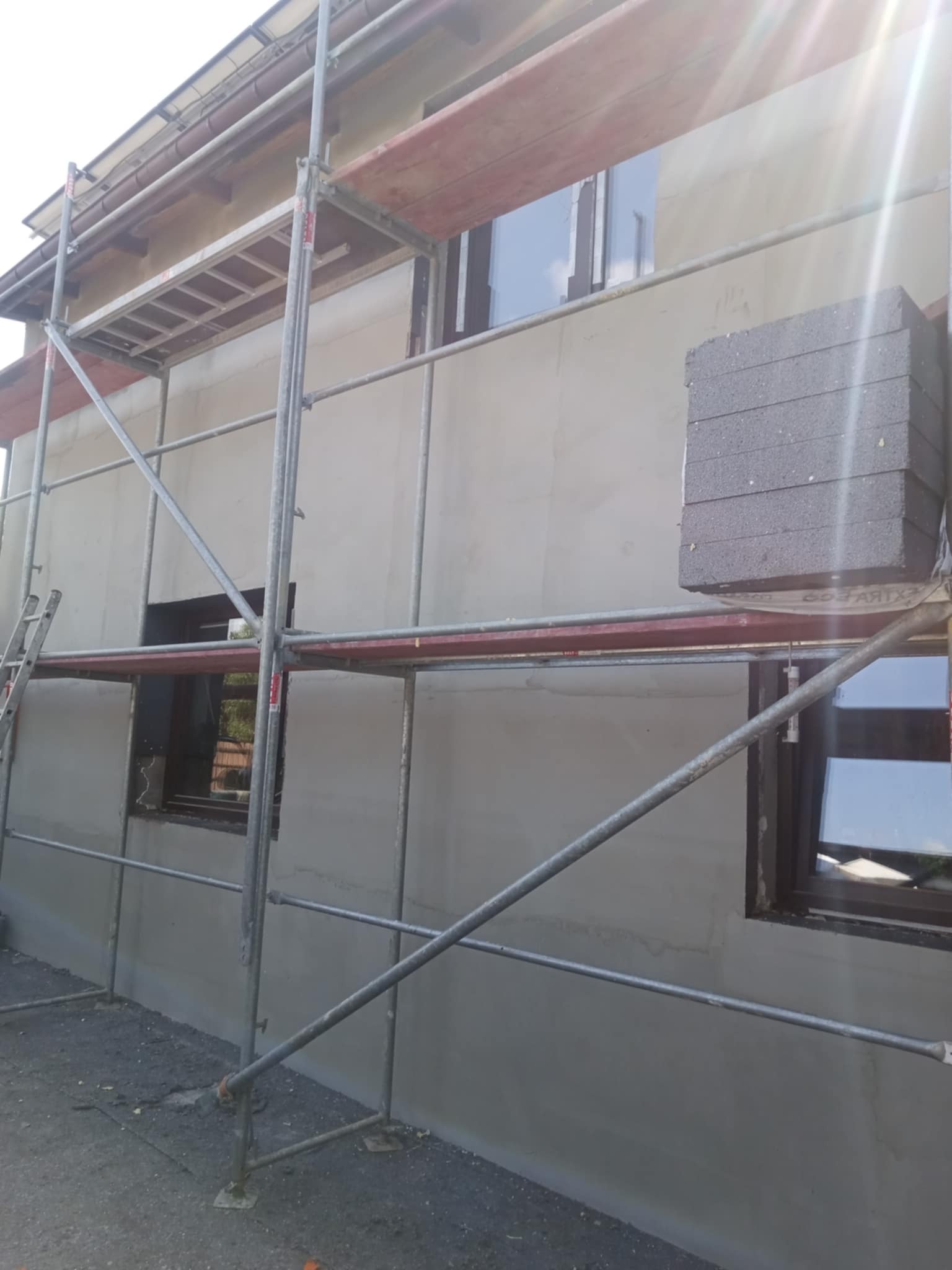 Ściana budynku w trakcie ocieplania, z widocznym rusztowaniem i styropianem. Świeżo nałożona warstwa kleju na elewacji. Okna z ciemną stolarką.