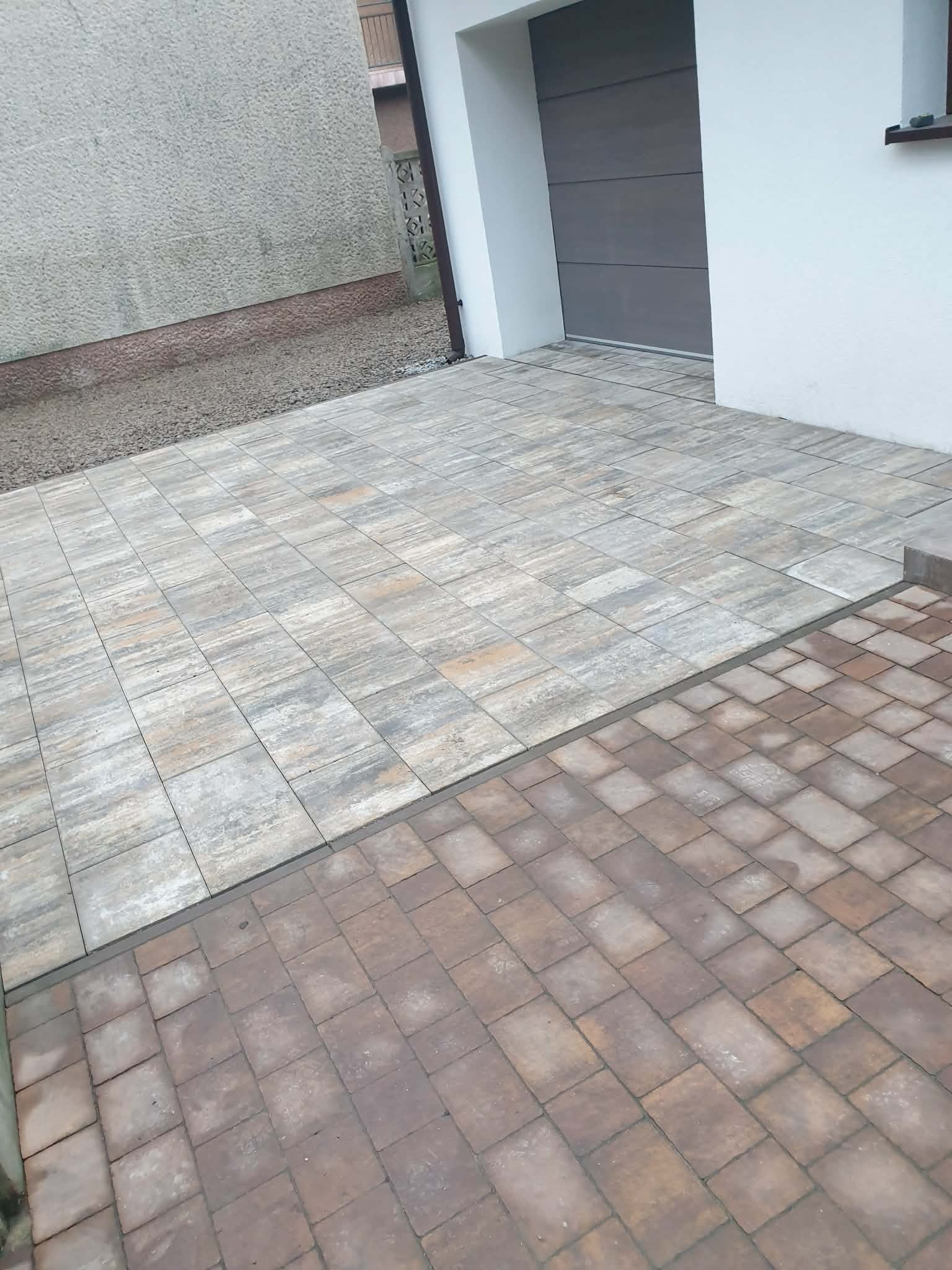 Ułożona kostka brukowa w dwóch odcieniach szarości i brązu przed garażem. Widoczna faktura kostki i staranne ułożenie. Szara ściana budynku w tle.