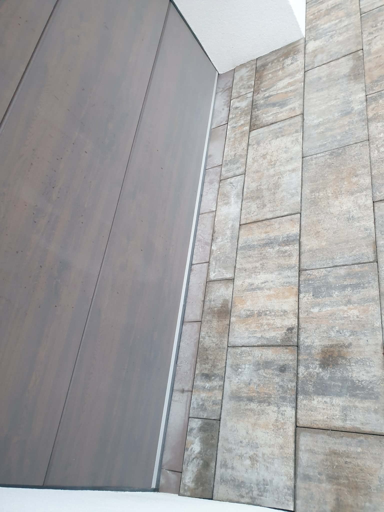 Nowoczesne wykończenie ściany z płytek imitujących kamień w odcieniach szarości i beżu, połączone z gładką, szarą powierzchnią. Widoczne detale łączeń i faktura materiałów.