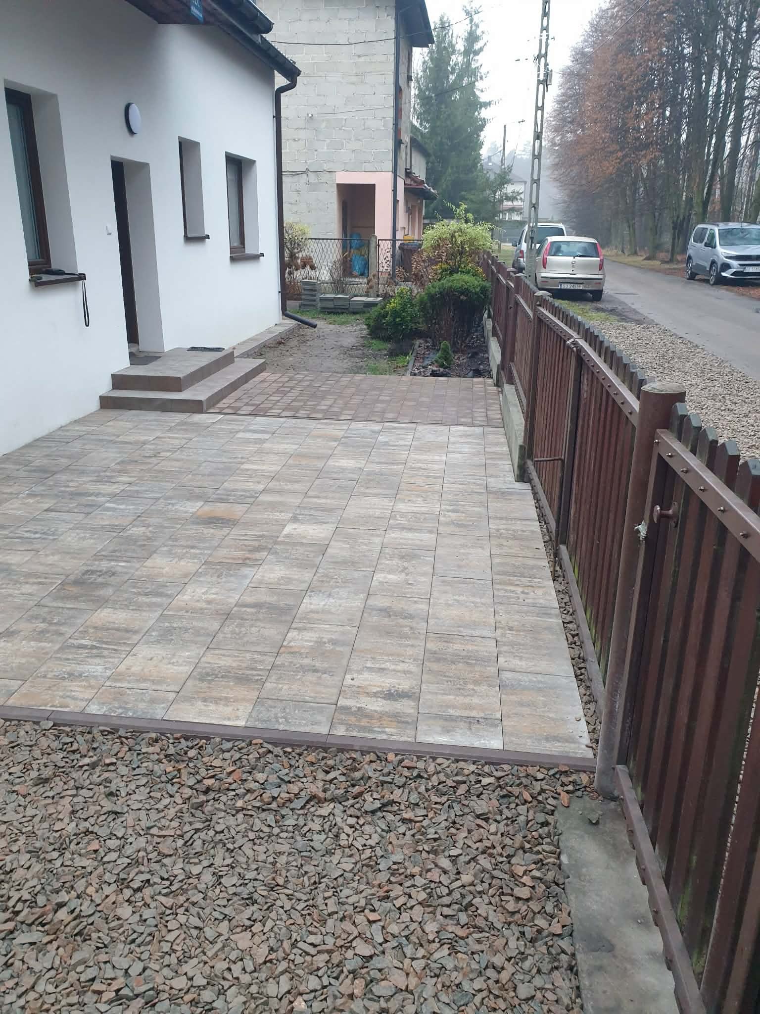 Ułożona kostka brukowa przed domem jednorodzinnym, widok z góry. Brązowe ogrodzenie po prawej stronie. Szara kostka brukowa ułożona w regularny wzór.