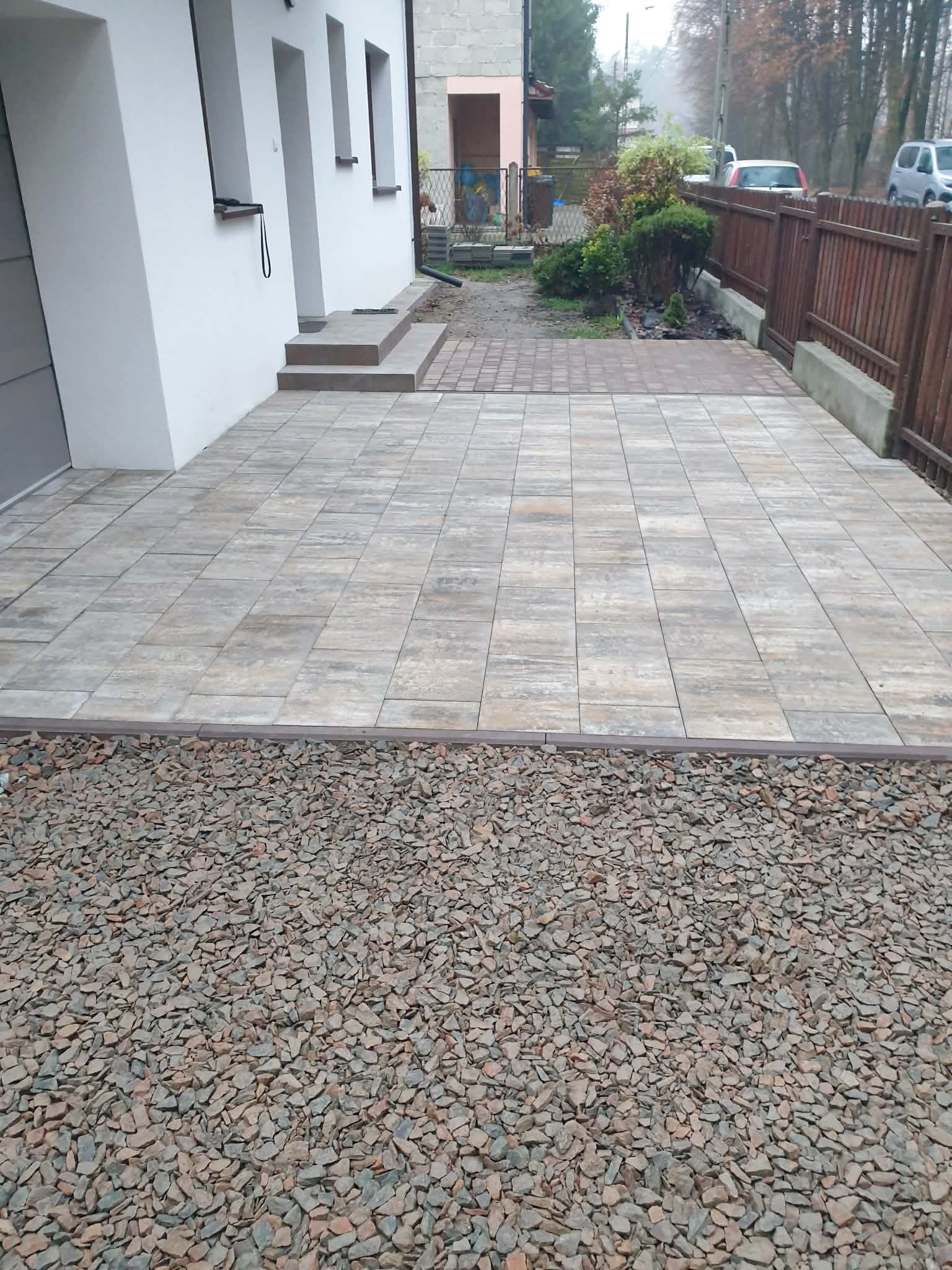 Ułożona kostka brukowa na podjeździe z obrzeżem i kamieniami ozdobnymi. Widok na dom z białymi ścianami i brązowym ogrodzeniem. Całość w stonowanej kolorystyce.
