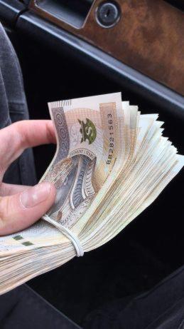 Plik banknotów polskich złotych, spięty gumką recepturką, trzymany w dłoni na tle wnętrza samochodu.