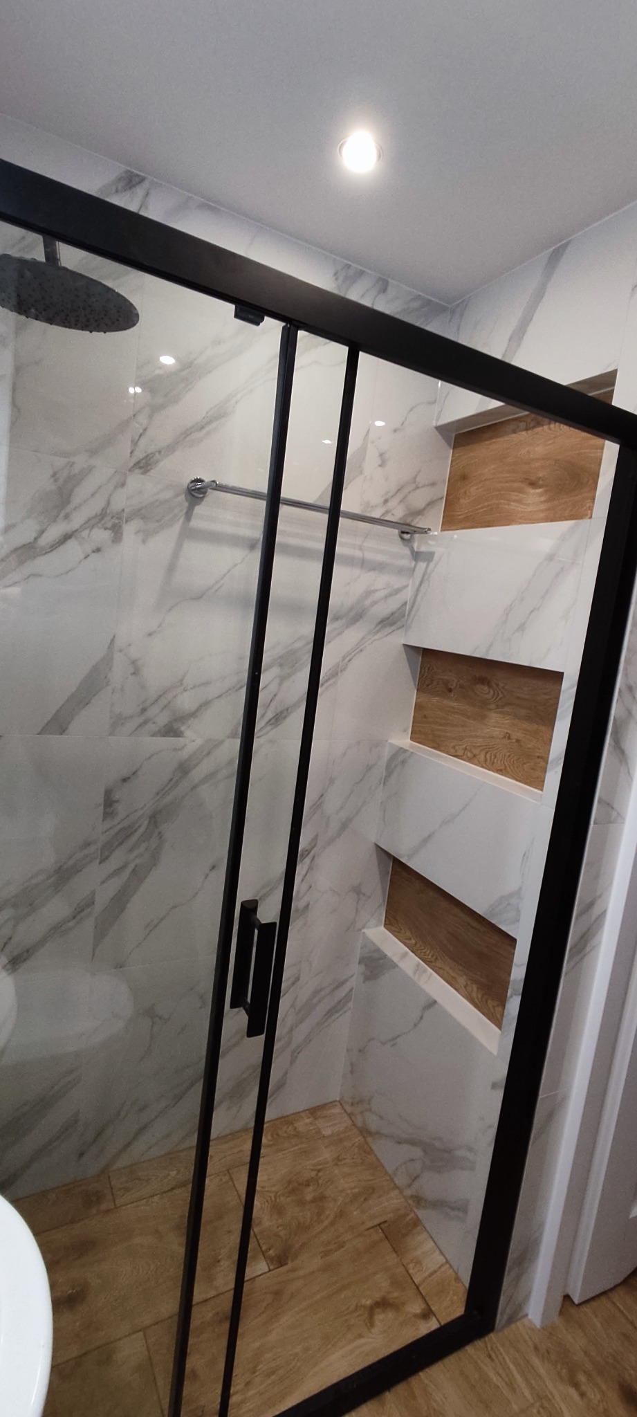 Nowoczesna kabina prysznicowa z czarną ramą, marmurowe płytki ścienne z drewnianymi wnękami, deszczownica i drewnopodobna podłoga.