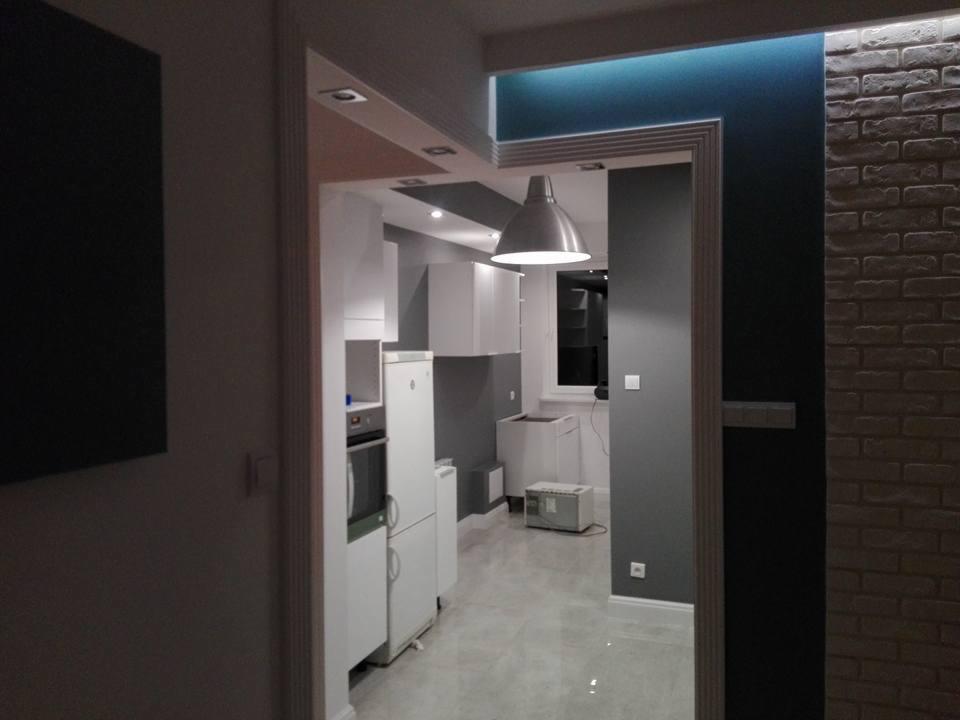 Widok z korytarza na nowocześnie wykończoną kuchnię z białymi meblami, szarą ścianą i ceglaną okładziną po prawej stronie, oświetloną lampą wiszącą i podświetleniem LED.