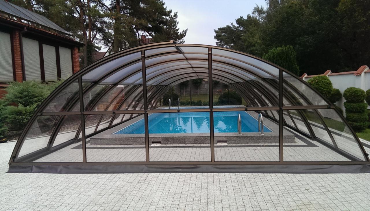 Zadaszony basen z błękitną wodą, otoczony szarą kostką brukową, widoczny z zewnątrz przez transparentne panele zadaszenia, na tle zieleni drzew i krzewów.