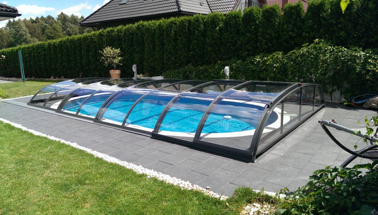 Zadaszony basen ogrodowy z błękitną wodą, otoczony szarym brukiem i zielonym trawnikiem, w tle rząd tuj.