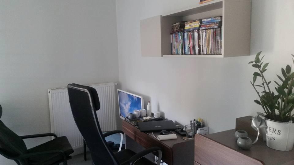 Gabinet z biurkiem, komputerem, dwoma krzesłami, półką z książkami i kwiatem doniczkowym, utrzymany w stonowanej kolorystyce.