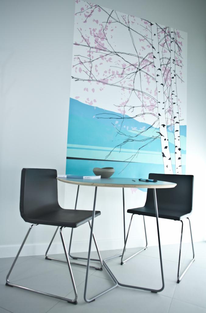 Minimalistyczna aranżacja wnętrza z okrągłym stołem, czarnymi krzesłami na metalowych nogach i abstrakcyjnym obrazem z motywem brzozy i różowych kwiatów na ścianie.