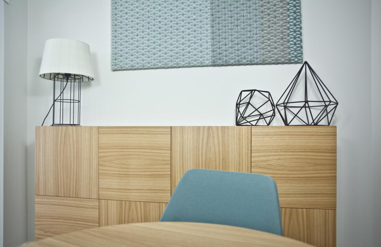 Nowoczesne wnętrze z drewnianą komodą, lampą z drucianym abażurem, geometrycznymi dekoracjami i abstrakcyjnym obrazem w odcieniach szarości i błękitu.