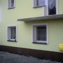 Jasnożółta elewacja domu z białymi obramowaniami okien, brązowym cokołem i żółtą skrzynką gazową, sfotografowana w pochmurny dzień.